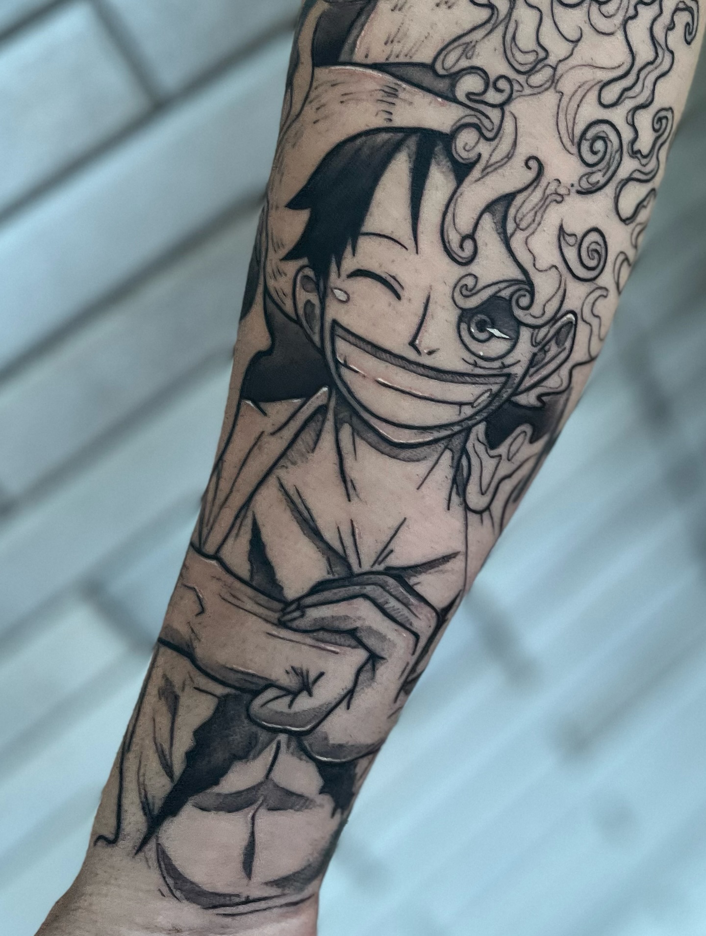 Tatuaje en el antebrazo que representa a un personaje de anime con una sonrisa amplia y detalles de humo, en un estilo artístico en blanco y negro.