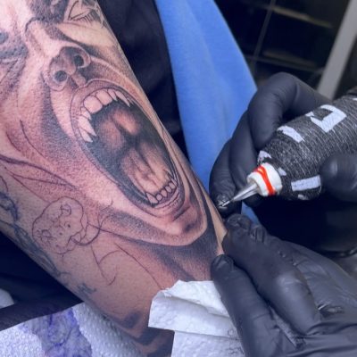 Tatuador trabajando en un tatuaje realista de un rostro gritando en el brazo de un cliente, destacando detalles y técnica de sombreado.