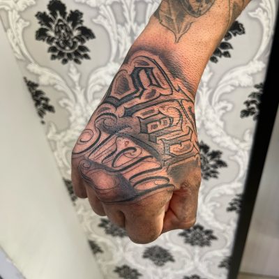 Tatuaje en la mano de un individuo, con diseño intrincado y detalles en negro, sobre un fondo con patrón decorativo.