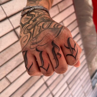 Tatuaje en la mano con la palabra "LIFE" en letras estilizadas, mostrando detalles de arte corporal en un fondo de ladrillos.