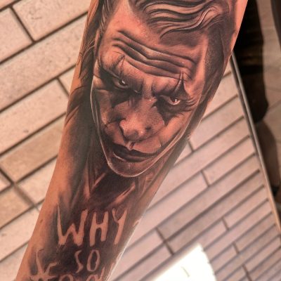 Tatuaje realista del Joker en el antebrazo, con el texto "WHY SO SERIOUS?" visible, sobre un fondo de ladrillos.